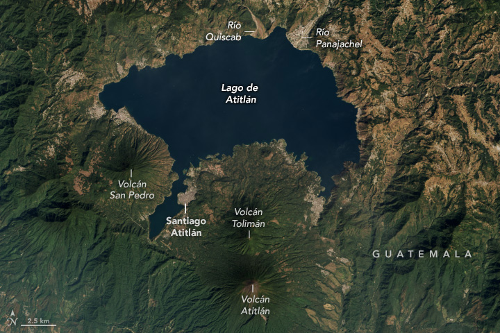 A dark-blue lake with an irregular coastline contains several wisps of faint green. Three prominent lush volcanoes (green) are at the image bottom, while patches of brown agricultural areas dot the middle and top.