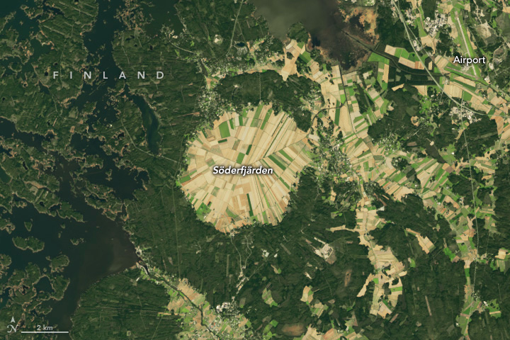 An impact crater appears as a hexagonally shaped area divided into many light green and tan agricultural fields in a roughly radial pattern. The fields contrast with darker green vegetation surrounding it. The left side and top of the image show some inlets of the Gulf of Bothnia.