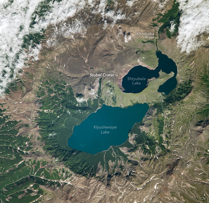 A top-down satellite image shows a dark blue lake and a lighter blue lake filling depressions in the ground within several overlapping calderas. The volcanic landscape is mostly brown, punctuated with green vegetation, especially around the lakes. Several valleys contain a thin white line of snow.