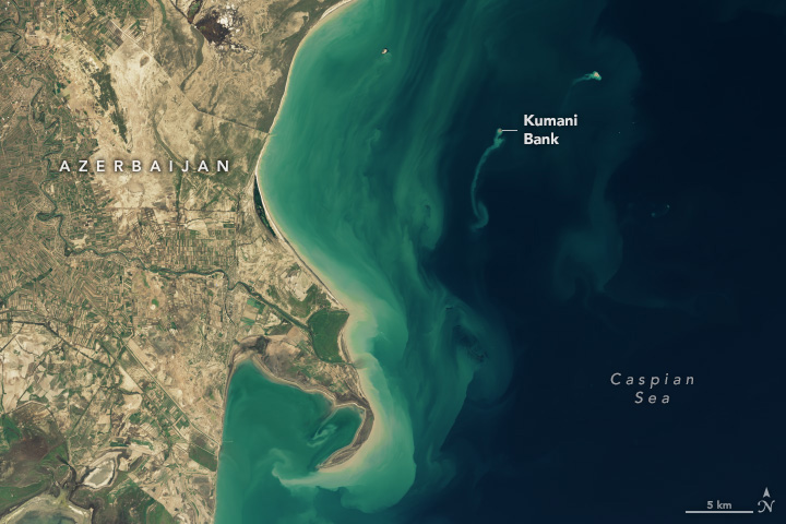 A satellite image showing a coastal area of Azerbaijan on the left and the dark blue Caspian Sea on the right. Coastal waters are cloudy with sediment, and the Kumani Bank mud volcano appears as a speck in the sea with a sediment plume streaming south from it.