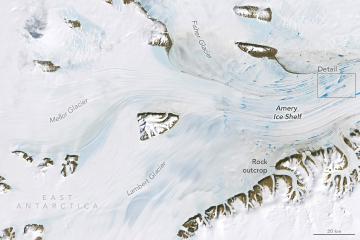 A downward-looking picture shows white ice flowing around and between several brown rock formations. The ice feeds into an ice shelf on the right side of the image, which is speckled with blue patches.