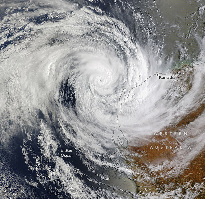 A downward-looking image shows swirling bands of white clouds around the eye of a tropical cyclone over blue ocean water. The cloud bands partially cover the coastal areas of Western Australia.