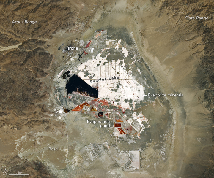 A labeled satellite image shows Searles Lake on August 15, 2024. The lake is mostly dry except for evaporation ponds with red and black fluid that are part of mining operations. A white crust of salty evaporite minerals is visible across much of the lake bed. Mountain ranges flank the lake to the east and west.