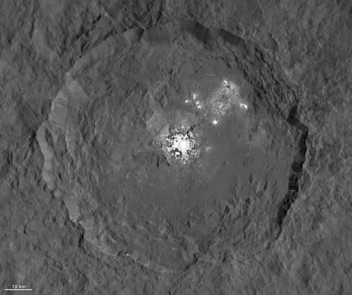 This satellite image from the Dawn spacecraft shows a large crater on the dwarf planet Ceres with a bright deposit in the middle of the crater. The deposit is an evaporite mineral composed of sodium carbonate. The image is black and white.