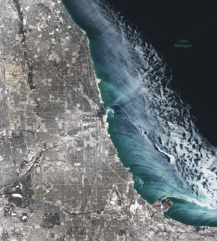 A downward-looking image shows gray and white urban areas on the left half of the scene. The right half of the scene shows dark blue water that is partially covered with lake ice, which transitions from dark to bright white.