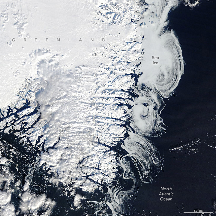 A satellite image shows the southern part of Greenland covered with white snow and ice. To the right of the island, smooth, white swirls of sea ice top the ocean, bordered to the right by dark, nearly black areas of open water.