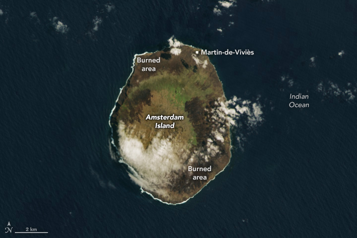 A natural-color satellite image is centered on an oval-shaped island in the dark-blue Indian Ocean. Much of the perimeter of the island is dark brown from being recently burned, while the rest is light green and light brown. A cloud obscures part of it.