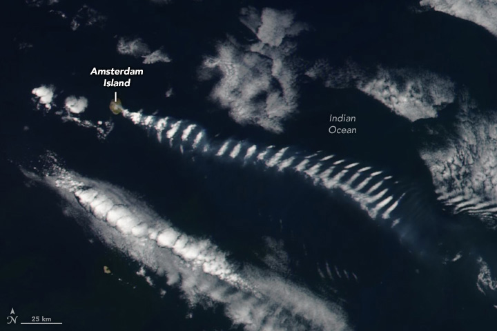 A satellite image shows an area of the Indian Ocean several hundred kilometers wide. Amsterdam Island is small, near the left side of the image. A trail of rippling clouds, along with a faint plume of wildfire smoke, extends to the right.