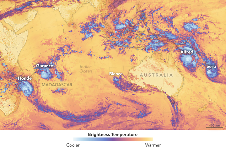 A false-color satellite image covering the southern Indian Ocean, Australia, and part of the South Pacific shows infrared signals called brightness temperature. Cooler signals in white and purple highlight five tropical cyclones: two near Madagascar, one off of Western Australia, and two east of Australia. They stand out from the rest of the image, which mostly shows warmer temperatures in yellow and orange.