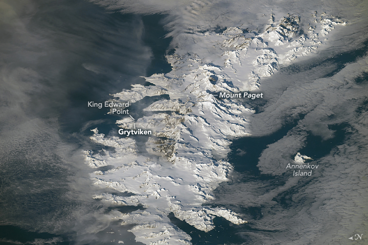 A long, narrow island spans the middle of a downward-looking photo. White snow and ice cover most of the island, which displays mountainous terrain down its spine. Dark ocean water and white clouds frame the island's left and right sides.