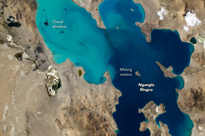 A nadir photo shows a vivid blue lake amid a brown, rocky landscape. The lake displays an irregular coastline and contains several islands. Its water is milky blue on the left side and transitions to a darker blue on the right side.